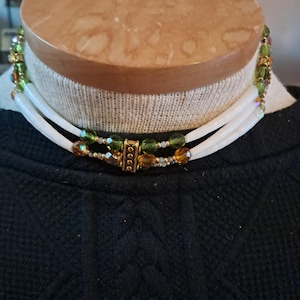 May include: A layered necklace with white, tube-shaped strands. The necklace features green and amber-colored beads, with gold-colored accents. The necklace is displayed on a mannequin head, showcasing its design and style.