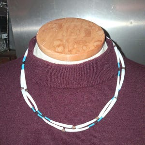 May include: A white necklace with a double strand design. The necklace features small turquoise and black beads, along with small metal accents. The necklace is displayed on a maroon sweater.