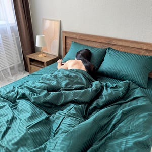 Könnte beinhalten: Ein Bett mit tealfarbener Bettwäsche, einschließlich Bettbezug und zwei Kissen. Die Bettwäsche hat ein Streifenmuster. Ein hölzernes Kopfteil ist sichtbar. Eine Person liegt im Bett und ist von der Bettdecke bedeckt.