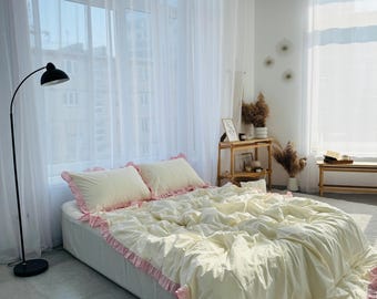 May include: A white bed with a ruffled pink trim and a white duvet. The bed is in a bedroom with white curtains and a lamp.
