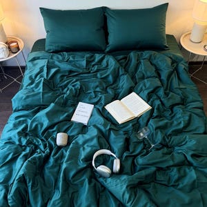 Könnte beinhalten: Ein Bett mit smaragdgrüner Bettwäsche, darunter zwei Kissen, eine Bettdecke und ein Spannbettlaken. Ein Buch, eine Kerze, ein Weinglas und weiße Kopfhörer liegen auf dem Bett. Zwei kleine Nachttische mit Lampen stehen auf beiden Seiten des Bettes.