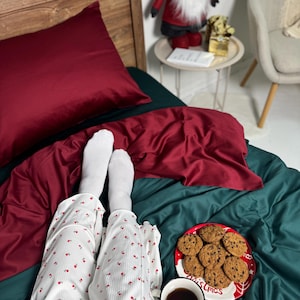 Könnte beinhalten: Gemütliche Schlafzimmerszene mit einem weinroten Kissen und einer Bettdecke auf einem teal-farbenen Bett. Ein Teller mit Schokoladen-Chip-Cookies und eine Tasse Kaffee liegen neben einem weißen Pyjama mit rotem Blumenmuster. Ein Weihnachtszwerg steht auf einem Beistelltisch.
