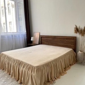 May include: A bed with a wooden headboard and a beige bed skirt. The bed is covered with a beige sheet. The room has a window with white curtains and a brown curtain. A small lamp and a framed picture are on a side table.