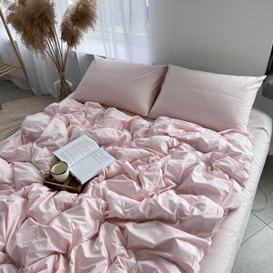 Könnte beinhalten: Ein rosafarbener Bettbezug mit einem Buch und einer Tasse darauf. Der Bezug liegt auf einem Bett mit zwei rosafarbenen Kissen.