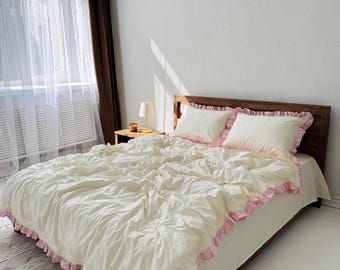 May include: A bed dressed with a cream-coloured duvet cover and two matching pillows. The duvet cover and pillowcases have a pink ruffled trim. The bed is against a white wall with a window and brown curtains. A small wooden bedside table with a lamp is next to the bed.