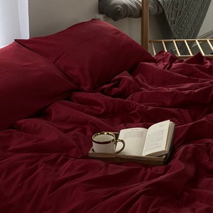 May include: A burgundy bed with two pillows and a book and a white mug on a wooden tray on top of the bedding.