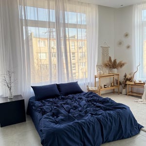Puede incluir: Una funda nórdica azul marino con textura arrugada, colocada sobre una cama con sábanas blancas y dos almohadas. La cama está en un dormitorio con paredes blancas y cortinas blancas transparentes.