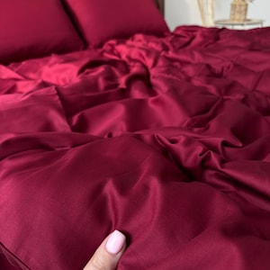 Puede incluir: Un juego de cama burdeos intenso, que incluye dos fundas de almohada y una sábana bajera, se muestra sobre una estructura de cama de madera. La ropa de cama tiene una textura sedosa y suave. La imagen muestra la ropa de cama con una mano sosteniendo la sábana, destacando la suavidad de la tela.