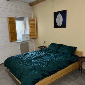 Könnte beinhalten: Ein Schlafzimmer mit einem Holzbettgestell und einer dunkelgrünen Bettdecke. Das Bett steht an einer weißen Wand mit Holzbalken an der Decke. Es gibt ein Fenster mit Holzläden und ein Bild an der Wand über dem Bett.