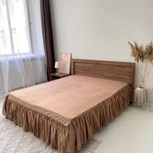 May include: A bed with a wooden headboard and a tan bed skirt. The bed is covered with a matching tan sheet. The room has a window with sheer curtains and a rug with a geometric pattern.