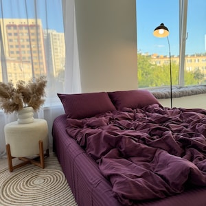 May include: A purple striped bedspread with a white pillow and a white side table with a plant on top. The bed is in a room with a window and a floor lamp.