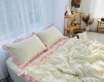 May include: A white bed with a white duvet and two white pillows with pink ruffled edges. The bed is made with a white bed skirt and has a pink ruffled trim. The bed is in a bedroom with white curtains and a wooden side table.