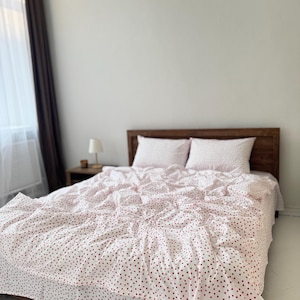 Op de afbeelding: Een bed met een houten hoofdbord en een witte dekbed- en kussenset. Het beddengoed is bedrukt met kleine rode hartjes. Een kleine lamp staat op een houten nachtkastje.