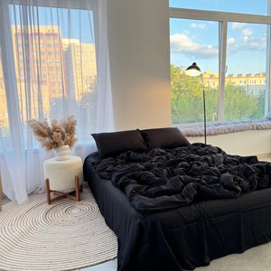 May include: A black bed with a white fluffy rug in front of it. The bed has a black comforter and two black pillows. There is a white vase with dried flowers on a white stool next to the bed.