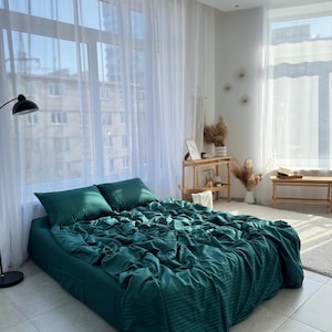 Könnte beinhalten: Ein grünes, gestreiftes Bettlaken mit weißen Streifen auf einem Bett mit weißen Kissen in einem Schlafzimmer mit großen Fenstern und weißen Vorhängen.