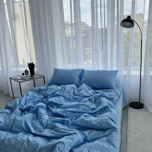 May include: A blue bed with white sheets and pillows. The bed is in a room with white curtains and a black floor lamp.