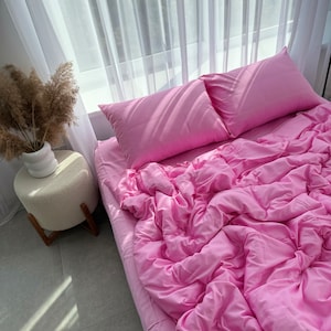 Puede incluir: Una cama con ropa de cama rosa, que incluye una sábana, un edredón y dos almohadas. La ropa de cama es de color rosa sólido y vibrante. La cama está cerca de una ventana con cortinas blancas transparentes. Un pequeño otomán redondo y un jarrón con plantas secas son visibles.