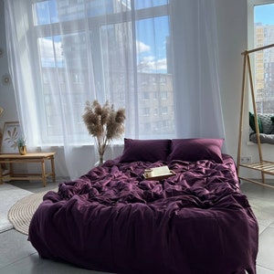 May include: A deep purple bedspread with white pillowcases and a book on a wooden tray. The bed is in a room with white walls and large windows with white curtains.