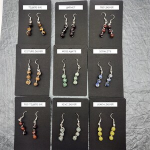 May include: Nine pairs of gemstone earrings displayed on black cards. Each card has the name of the gemstone written on it. The earrings are made with silver hooks and three gemstone beads.