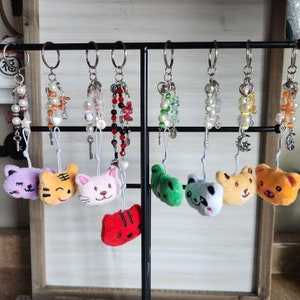May include: Nine colorful beaded keychains with plush animal charms. The charms include a purple cat, orange tiger, pink cat, green frog, white panda, yellow bear, and orange bear. The keychains are hanging on a black metal rack.