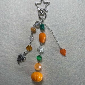 May include: A silver keychain with a star-shaped clasp, adorned with a charm featuring a pumpkin, leaf, and various beads in orange, green, and clear colors. The charm is attached to a chain.