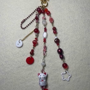 May include: A decorative keychain featuring a gold-toned star clasp with three dangling beaded strands. The strands include red and white beads, a white cat charm, a red square button, a white round button, and a small white open star charm.