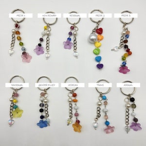 May include: Keychains with various Pride flag color schemes. Each keychain features a silver ring, decorative beads, chain, and a flower charm. Text labels include "Pride 1", "Non Binary", "Bisexual", "Pride 2", "Pride 3", "Pansexual", "Gender Fluid", "Lesbian", "Trans", and "Asexual".