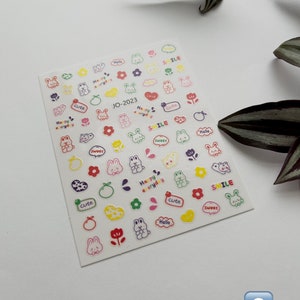 May include: A sheet of colorful nail stickers featuring cute cartoon bunnies, flowers, hearts, and oranges. The stickers have various phrases including "Happy everyday", "Smile", "Cute", "Sweet", "Love", and "Hello".