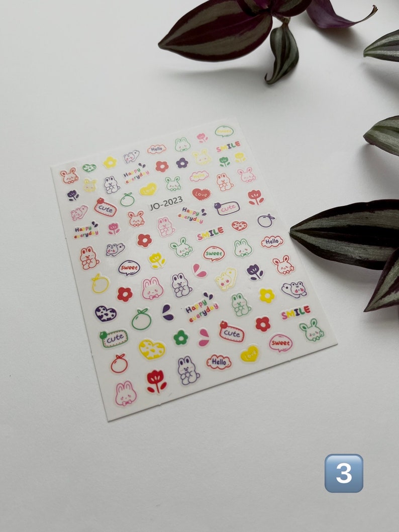 May include: A sheet of colorful nail stickers featuring cute cartoon bunnies, flowers, hearts, and oranges. The stickers have various phrases including "Happy everyday", "Smile", "Cute", "Sweet", "Love", and "Hello".