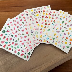 May include: Five sheets of colorful nail stickers with various designs, including watermelon slices, peaches, flowers, and cute cartoon animals. The sheets are labeled with the text "JO-2015", "JO-2012", "JO-20", and "JO-2020".