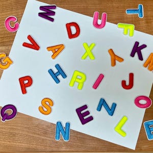 May include: A white sheet of paper with colorful felt letters scattered across it. The letters are in various colors, including red, orange, yellow, green, blue, purple, and pink. The letters are arranged in a random order, and some of them are overlapping.
