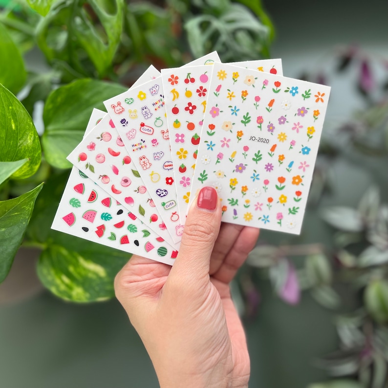May include: A set of six sheets of colorful nail stickers featuring various designs, including watermelon slices, peaches, cherries, flowers, and cute cartoon animals. The stickers are on white sheets with a white background.