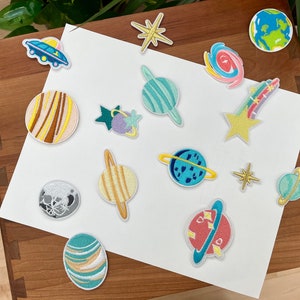 May include: A collection of colorful embroidered iron-on patches featuring planets, stars, and a spaceship. The patches are on a white background.