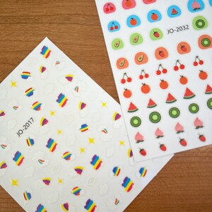 May include: Two sheets of nail stickers. One sheet features a rainbow and cloud design with the text "JO-2017". The other sheet features a variety of fruit designs, including watermelon, kiwi, strawberries, peaches, and cherries. The text "JO-2032" is on the sheet.