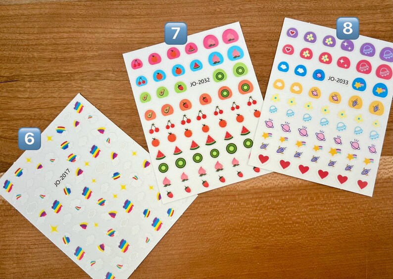 May include: Four sheets of colorful stickers with different designs. The first sheet has rainbow-colored shapes, the second sheet has fruit designs, the third sheet has fruit designs, and the fourth sheet has space-themed designs. The sheets are labeled with the numbers 6, 7, and 8.