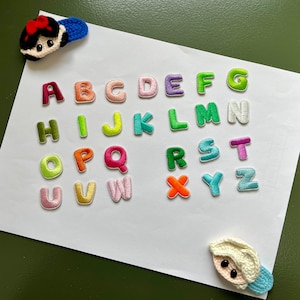 May include: A white sheet of paper with colorful felt letters of the alphabet, A through Z, arranged in rows. There are two small felt figures, one of a girl with a red bow and one of a girl with a white bow, on the paper.