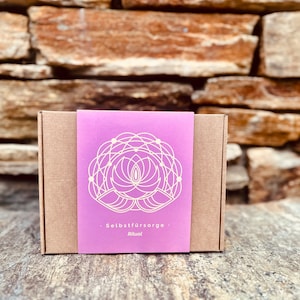 Op de afbeelding: Een bruine kartonnen doos met een roze deksel. Het deksel heeft een wit geometrisch ontwerp en de tekst "Selbstfürsorge Ritual" in wit.