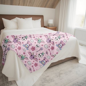 K&ouml;nnte beinhalten: Eine rosa Fleece-Decke mit einem sich wiederholenden Muster aus niedlichen Cartoon-Flederm&auml;usen, Sch&auml;deln, Blumen und Wolken. Die Flederm&auml;use sind lila und gr&uuml;n mit rosa Fl&uuml;geln. Die Sch&auml;del sind wei&szlig; mit rosa Akzenten. Die Blumen sind rosa und wei&szlig;. Die Wolken sind wei&szlig;.
