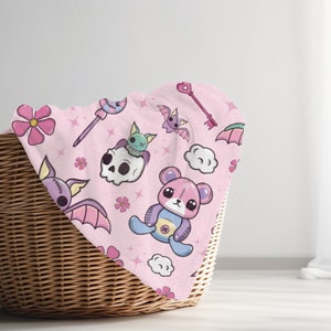 K&ouml;nnte beinhalten: Ein rosa Stoff mit einem sich wiederholenden Muster aus niedlichen Comicfiguren, darunter Flederm&auml;use, ein Sch&auml;del, ein Teddyb&auml;r, Blumen und Sterne. Der Stoff ist gefaltet und in einem Weidenkorb gelegt.