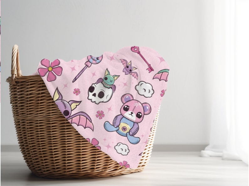 K&ouml;nnte beinhalten: Ein rosa Stoff mit einem sich wiederholenden Muster aus niedlichen Comicfiguren, darunter Flederm&auml;use, ein Sch&auml;del, ein Teddyb&auml;r, Blumen und Sterne. Der Stoff ist gefaltet und in einem Weidenkorb gelegt.