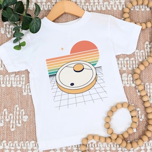 May include: White t-shirt with a retro-style graphic of a robot vacuum cleaner on a grid pattern with a rainbow sunset in the background.