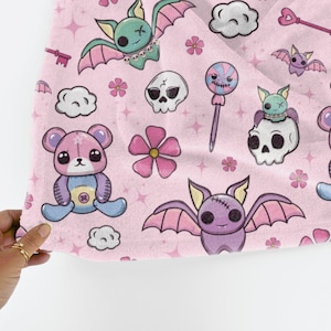 K&ouml;nnte beinhalten: Rosafarbener Stoff mit einem sich wiederholenden Muster aus niedlichen Cartoon-Flederm&auml;usen, Teddyb&auml;ren, Sch&auml;deln, Blumen und S&uuml;&szlig;igkeiten. Der Stoff hat eine weiche, flauschige Textur.