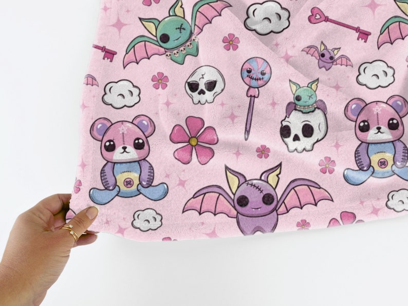 K&ouml;nnte beinhalten: Rosafarbener Stoff mit einem sich wiederholenden Muster aus niedlichen Cartoon-Flederm&auml;usen, Teddyb&auml;ren, Sch&auml;deln, Blumen und S&uuml;&szlig;igkeiten. Der Stoff hat eine weiche, flauschige Textur.