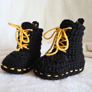 May include: A pair of black crochet baby boots, resembling work boots, with yellow laces and stitching. The handmade boots have a textured finish and a soft appearance. The boots are designed for a baby.