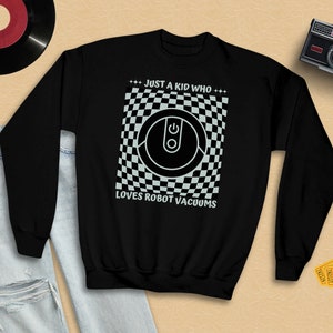 Può includere: Felpa nera con una grafica bianca di un aspirapolvere robot su uno sfondo a scacchi. Il testo "*** JUST A KID WHO *** LOVES ROBOT VACUUMS" è stampato sotto la grafica.