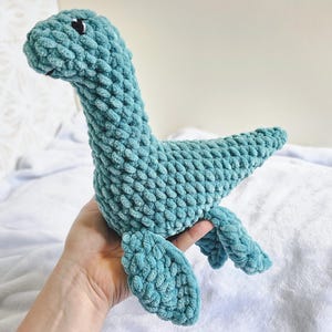 May include: A teal-coloured, plush Loch Ness Monster toy. The handmade stuffed animal has a long neck, small black eyes, and flipper-like appendages. The soft, textured material gives the toy a cuddly appearance.
