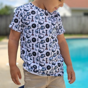 以下が含まれることがあります： 掃除用具や用品のカラフルなパターンがプリントされた白い半袖Tシャツ。プールの前で立っている子供が着用しています。