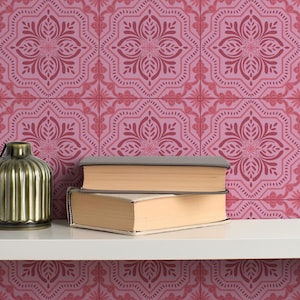 May include: A decorative shelf displays a gold-colored, ribbed vase and two books with beige pages and gray covers. The background features a pink floral tile pattern. The shelf is white.