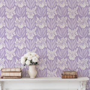May include: A lilac-colored floral wallpaper with a repeating pattern of stylized white irises. The wallpaper is the backdrop for a white mantelpiece, a vase of white flowers, and stacks of books.