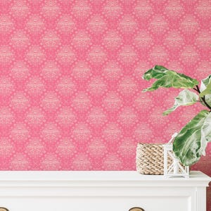 May include: Pink wallpaper with a repeating damask pattern. A white dresser and a green plant with large leaves are in the foreground. A small woven basket and a white decorative lantern sit on the dresser.